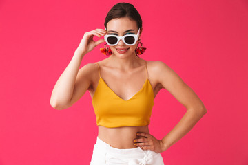 Photo of fashionable european woman 20s wearing fashion earrings touching sunglasses and smiling at you, isolated over pink background