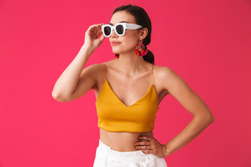 Photo of glamour caucasian woman 20s wearing fashion earrings touching sunglasses and looking aside, isolated over pink background