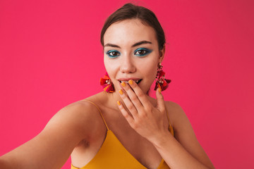 Photo of excited elegant woman 20s wearing earrings covering mouth with hand while taking selfie, isolated over pink background