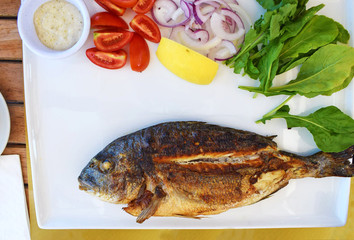Baked dorado fish on a white plate with greens, tomatoes, lemon, onion and sauce. Restaurant menu with fish and vegetables. Turkey. Grilled Dorade Royale Fish on the plate