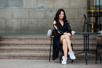 Young beautiful stylish girl goes in a black dress in the city. Outdoor summer portrait of a young woman with a hat