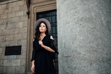 Young beautiful stylish girl goes in a black dress in the city. Outdoor summer portrait of a young woman with a hat