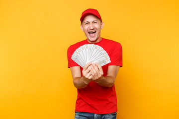 Delivery man in red uniform isolated on yellow orange background. Professional male employee in cap, t-shirt courier or dealer holding bundle lots of dollars, cash money. Service concept. Copy space.