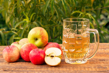 Glass goblet of apple cider and harvested apples on wooden boards with leaves of apple tree on the background