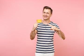 Portrait of smiling young man in striped t-shirt holding credit bank card, copy space isolated on trending pastel pink background. People sincere emotions lifestyle concept. Advertising area workspace