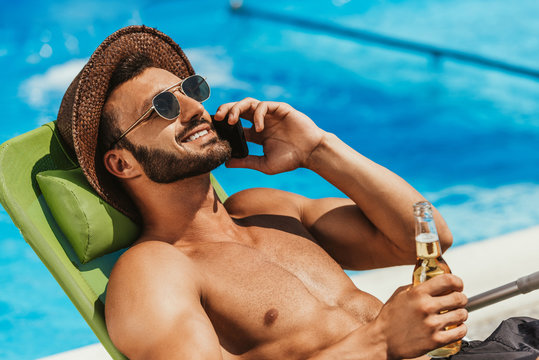 man in sunglasses talking on smartphone and holding bottle of beer on sunbed at poolside