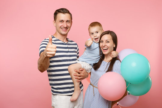 Portrait of young happy family, parents keep in arms, kissing hugging child kid son baby boy, celebrating birthday holiday party on pink background with colorful air balloons. Sincere emotions concept