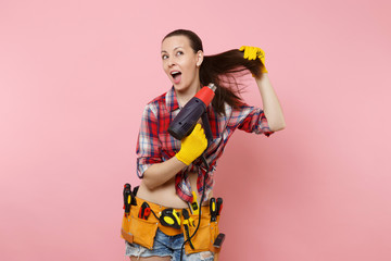 Young beautiful fun handyman woman in plaid shirt, denim shorts, kit tools belt full of variety instruments holding power heat gun isolated on pink background. Female in male work. Renovation concept.