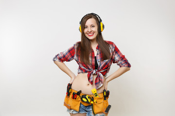 Handyman woman in shirt, denim shorts, noise insulated headphones, kit tools belt full of instruments standing with arms akimbo isolated on white background. Female in male work. Renovation concept.