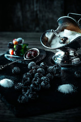 Still life in royal style. Frozen berry of blackberry with silver sugar bowl and spoon on black stone stand. Vertical shot