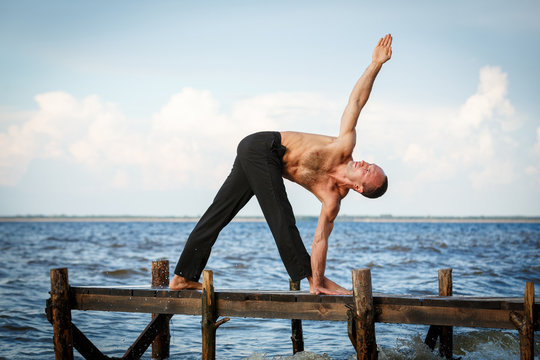 Young yoga trainer practicing parivrita trikonasana on a wooden pier on a sea or river shore