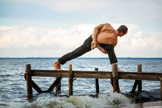 Young yoga trainer practicing parivrita Parswakonasana pose on a wooden pier on a sea or river shore