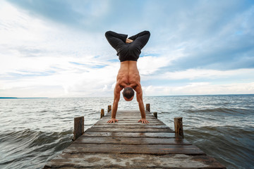 Young yoga trainer practicing handstand with a lotus on a wooden pier on a sea or river shore.