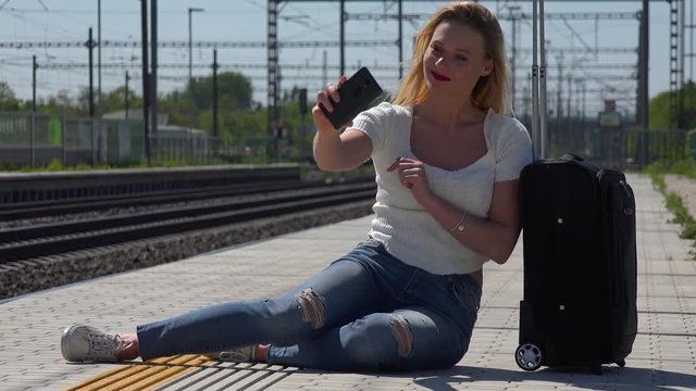 A young beautiful woman sits by a suitcase on a train station platform and takes selfies with a smartphone
