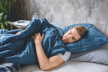 young handsome man sleeping under blanket in his bed at home