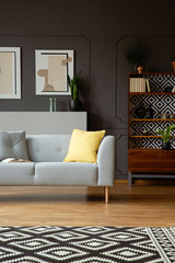 Yellow pillow on grey couch in retro living room interior with patterned carpet and posters. Real photo