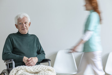 Sad disabled elderly man in green sweater in a hospital with blurred nurse
