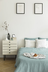 Real photo of white bedroom interior with breakfast placed on bed with pillows and pastel green bedclothes, two simple posters and bedside table with decor