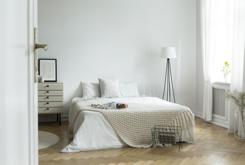 White bedroom interior in real photo with king-size bed with knit blanket and breakfast tray, window with curtains and empty wall with place for your poster - obrazy, fototapety, plakaty