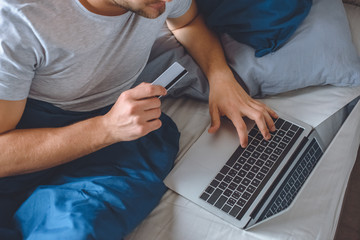 cropped image of man in bed with credit card doing online shopping on laptop