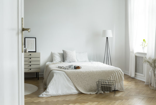 White bedroom interior in real photo with king-size bed with knit blanket and breakfast tray, window with curtains and empty wall with place for your poster