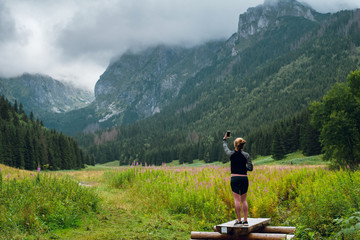 Rear view shot of young woman standing on the wooden dock on small pond and making photos of beautiful green hills and rocky peaks of Rysy mountains, Tatra. Searching for good phone signal