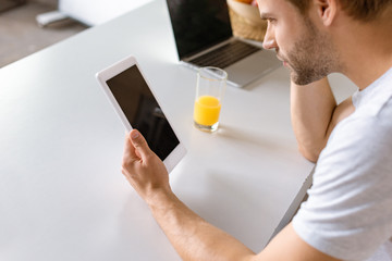 man using digital tablet with blank screen at kitchen table with laptop and glass of juice