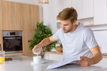 young man with newspaper pouring milk into cup with coffee at kitchen table