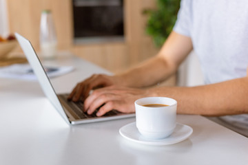 selective focus of coffee cup and male using laptop at kitchen table