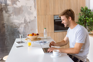 smiling young man using laptop at kitchen table with digital devices and coffee cup