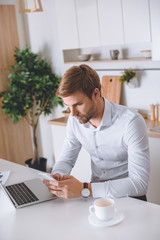 serious young businessman using smartphone during breakfast at kitchen table with laptop and coffee