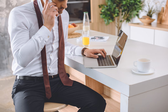 cropped image of businessman in white shirt with necktie over neck talking on smartphone and working on laptop at kitchen
