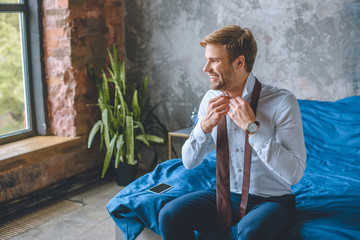 smiling businessman tying necktie in bedroom at home