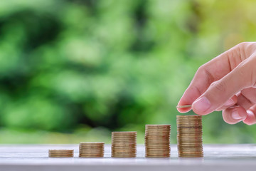 Woman hand putting coin, gold coins stack on wooden table in the morning sunlight. business,...