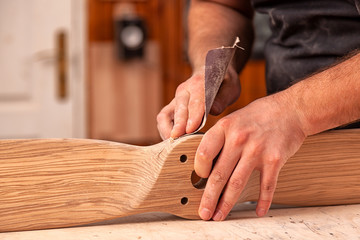 A working man in a  cap and shirt polishes the wooden block with sandpaper before painting in the workshop, in the background, tools and drilling machine.
