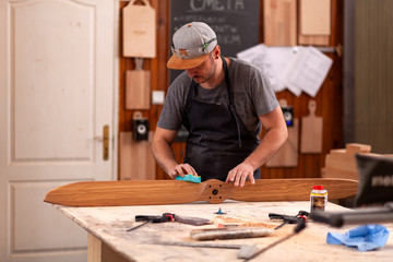 A working man in a  cap and shirt polishes the wooden block with sandpaper before painting in the workshop, in the background, tools and drilling machine.