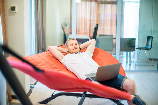 Young man with laptop holding hands behind head, relaxing and dreaming while lying on a hammock in office space. Taking time for a minute break. Unproductive working time. Selective focus. Copy space. - Powered by Adobe