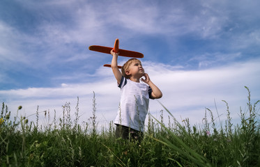 Little boy is launching a toy airplane in a field against a blue sky with clouds. He looks dreamily...
