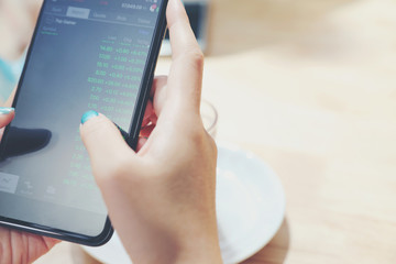 Close up on woman hand holding smartphone trading stock and data in coffee shop background business concept