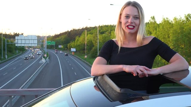 A young beautiful woman stands through a sunroof in a car and talks to the camera with a smile - a highway in the background