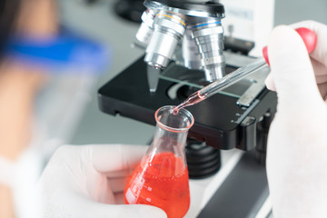 Hands of young Asian man and white microscope in science laboratory with red liquid and dropper for testing. Technician dripping red water on slide glass in science lab.
