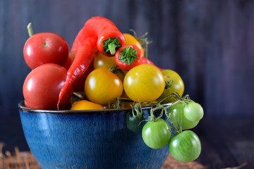 Cherry tomatoes of different colours and chili peppers in a blue bowl against the dark background