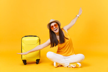 Tourist woman in summer clothes, hat sit at suitcase crossed legs spreading hands as in flight isolated on yellow orange background. Passenger traveling abroad on weekend getaway. Air journey concept.