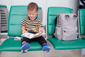 Fototapeta na wymiar Little traveler, cute smiling little Asian 30 months / 2 years old toddler boy child having fun reading a book while waiting for his flight at gate in terminal at airport, Traveling with kid concept