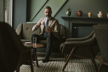 Handsome senior businessman drinking coffee and using mobile phone in lobby
