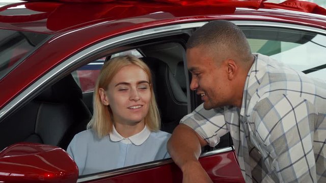 Beautiful young woman sitting in her new car, talking to her boyfriend. Happy couple smiling to the camera holding car keys, posing with their new auto. Consumerism, love, family concept.