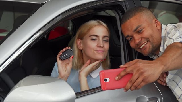 Beautiful young happy woman sitting in an auto holding car keys while her boyfriend using smart phone, taking selfies. Cheerful couple celebrating buying new automobile. Transport concept.