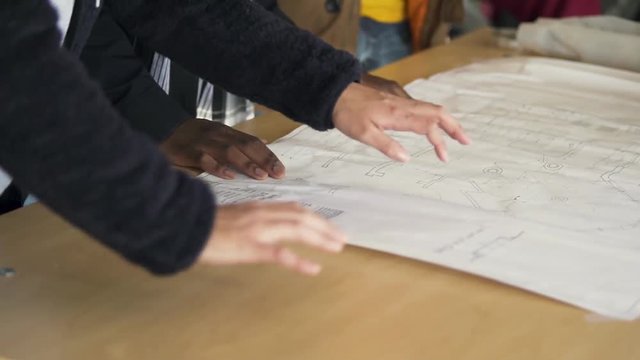Three young people discuss the plans for a building project in a work area. They gesture with their hands as they discuss.