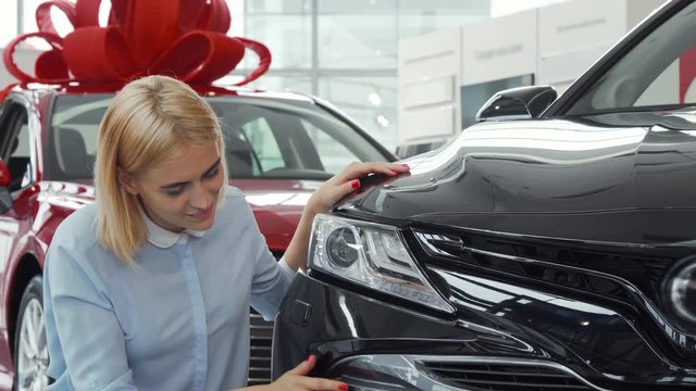 Cheerful young beautiful woman smiling joyfully to the camera while examining a new car for sale at the local dealership. Happy female customer posing with her newly bought auto.