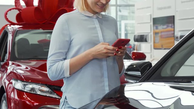 Cropped shot of a woman smiling, typing messages on her smart phone while buying a car at the cars showroom. Female customer surfing internet at the dealership. Technology, online internet concept.